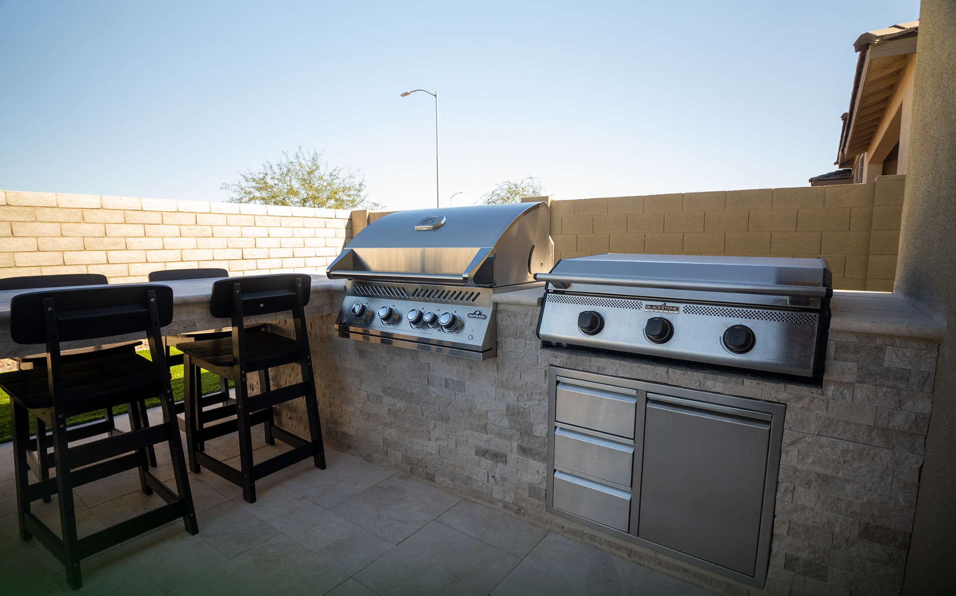 Outdoor kitchen with stainless grill and griddle behind a stone bar with black stools
