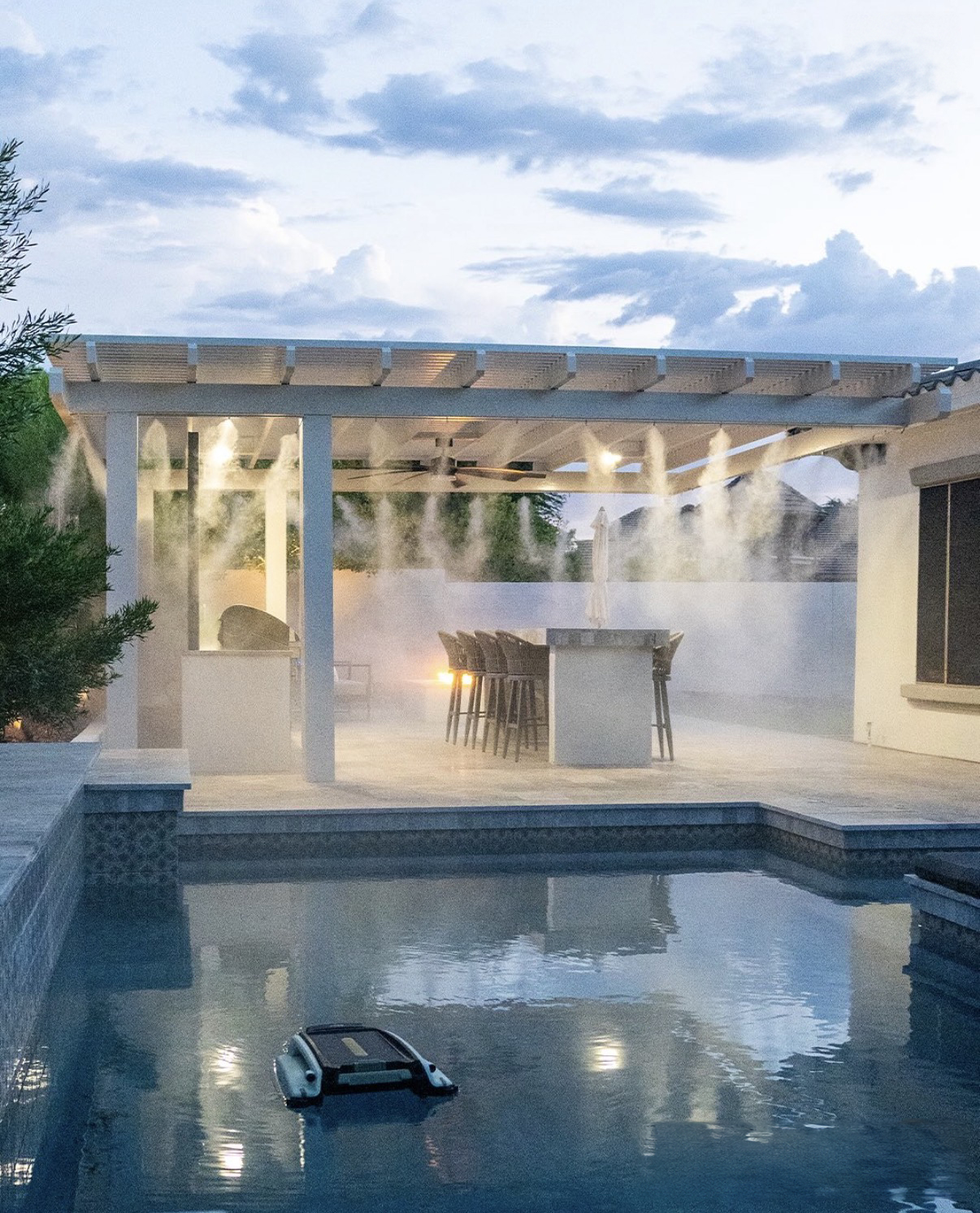 Pool at dusk with mist falling from a white pergola over bar seating and an outdoor kitchen