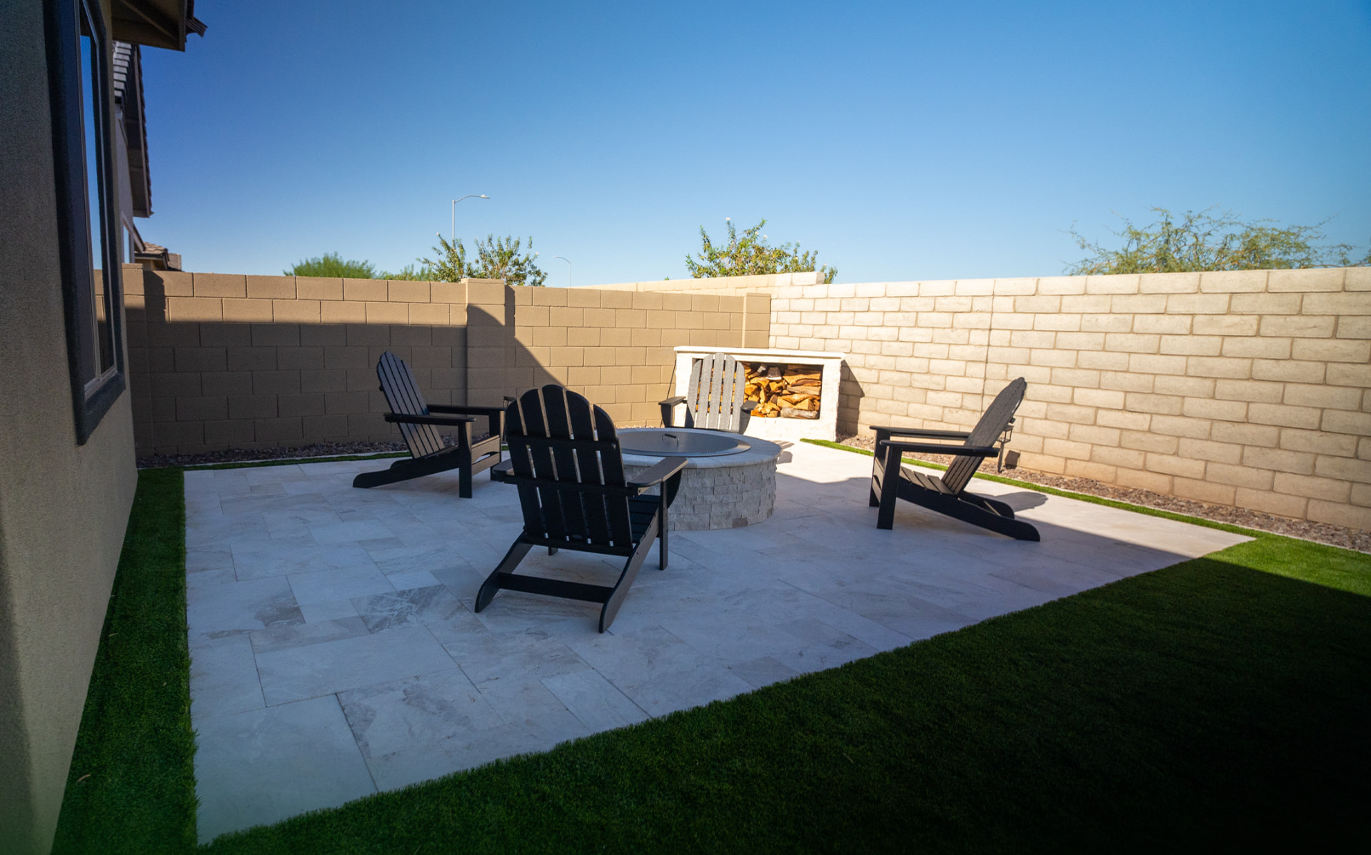 Round stacked-stone fire pit with four black Adirondack chairs on a travertine patio