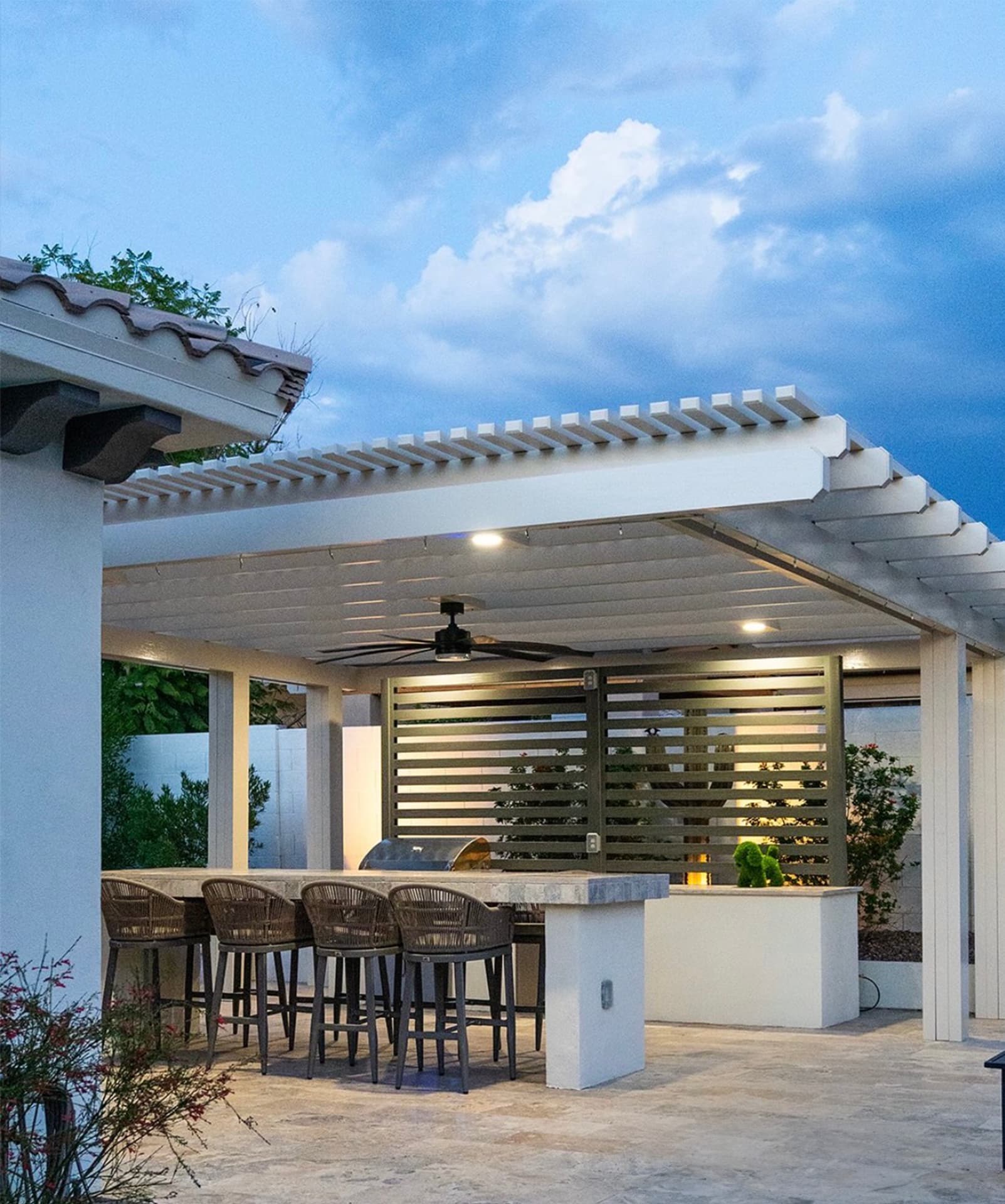 White louvered pergola over outdoor kitchen with stone bar, stools and olive slat wall
